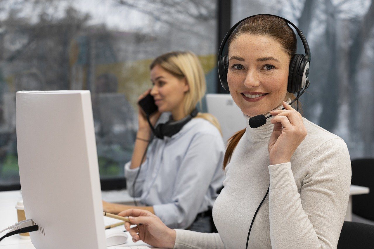 A woman wearing a headset and smiling, working at a computer in a call center, with a colleague in the background on the phone.